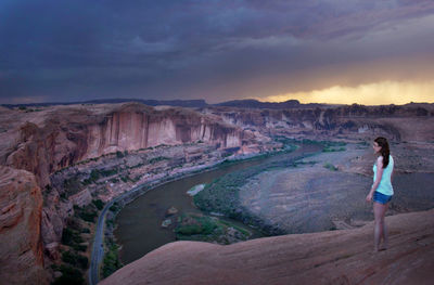 Side view of woman standing on cliff by river against cloudy sky during sunset