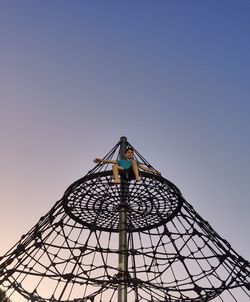 Low angle view of boy sitting on jungle gym against clear blue sky