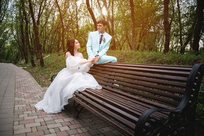 Full length of bride and groom sitting on bench in park