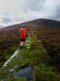 Rear view of backpack man walking towards mountain against cloudy sky