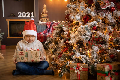 Cute boy holding gift box sitting by christmas tree at home