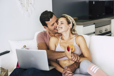 Smiling man and woman kissing while using laptop on sofa at home