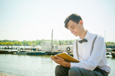 Young man writing in book while sitting at harbor against sky
