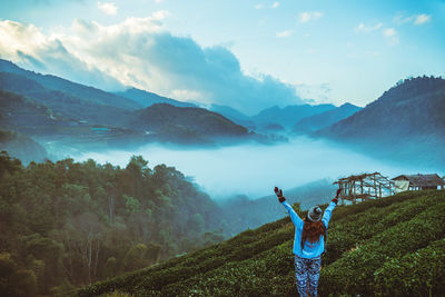 Rear view of woman with arms raised standing by plants against sky