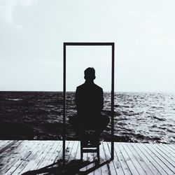 Rear view of silhouette man sitting on pier against river and sky during sunny day