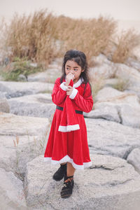 Girl wearing red dress holding santa hat while standing on rocks