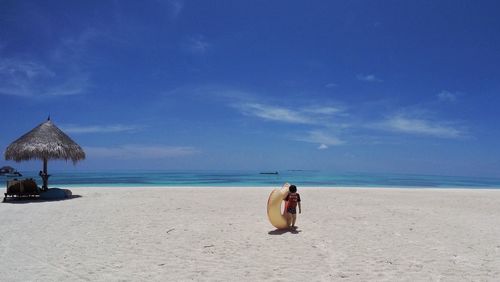 Full length of boy with inflatable ring walking at beach against blue sky during sunny day