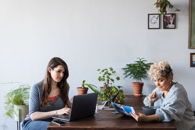 Woman using laptop while roommate reading newspaper at table against wall in living room