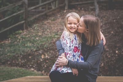 Mother kissing daughter while sitting on bench at park