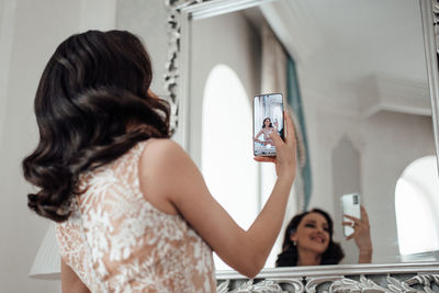 Rear view of woman using phone while standing in mirror