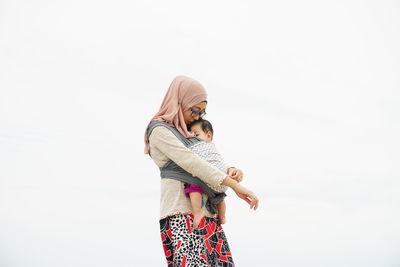 Mother wearing hijab carrying daughter against sky