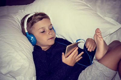 Cute boy listening to music on headphones while lying on bed at home