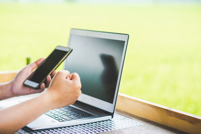 Cropped hands of woman using mobile phone by laptop on wooden table against landscape