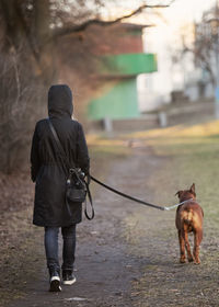 A girl in a black parka walks with a dog on the road to the distance, people from behind