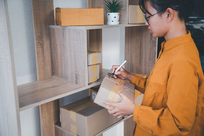 Girl writing over box while standing by shelf
