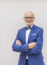 Portrait of smiling senior man in suit with arms crossed standing against white background