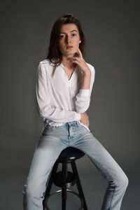 Portrait of young woman sitting on stool against gray background