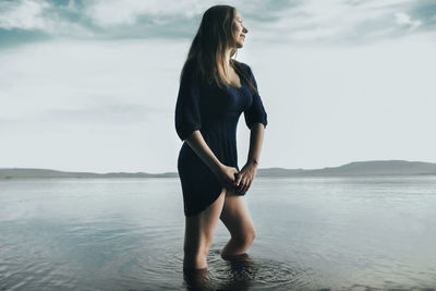 Thoughtful woman squeezing dress while standing in sea against sky