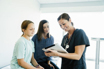 Cheerful pediatrician showing tablet computer to mother and daughter sitting on examination table in hospital