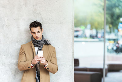 Young man using smart phone while standing against wall