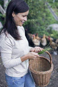 Smiling woman holding grass and wicker basket at poultry farm