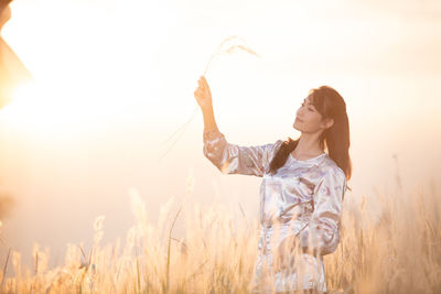 Mid adult woman holding plant while standing on field against sky during sunset