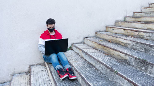Young caucasian man working on his laptop, while sitting on some stairs, outdoors, with a face mask