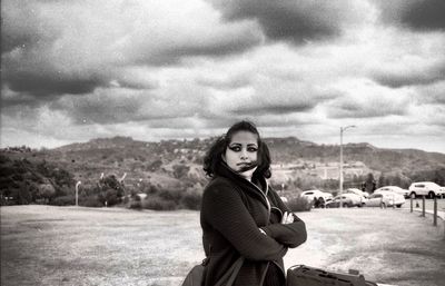 Thoughtful woman with arms crossed standing on field against cloudy sky