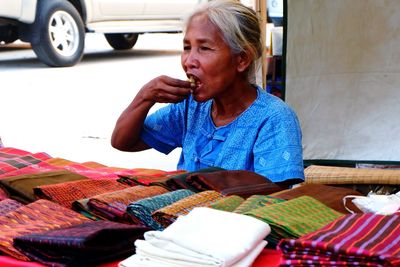 Senior woman eating betel nut while sitting by colorful fabrics at market