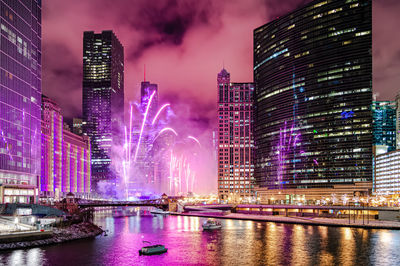 Illuminated modern buildings by river against sky at night