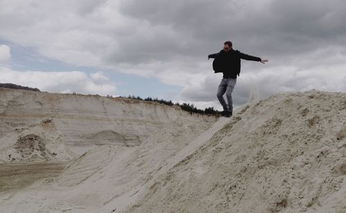 Happy man with arms outstretched jumping on sand against cloudy sky