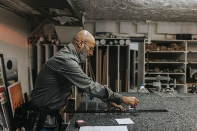 Mature craftsman measuring glass while standing at workbench