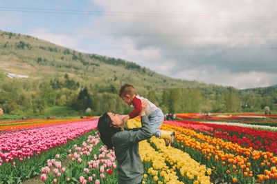 Side view of mother lifting son while standing by colorful flowering plants on field against sky