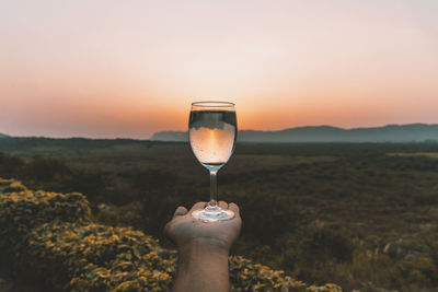 Person holding wineglass against sky during sunset
