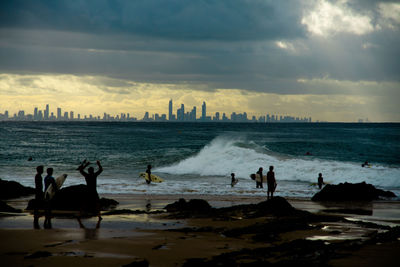 People enjoying at beach by city against cloudy sky during sunset