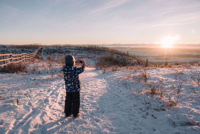 Young boy taking pictures in a snowy field at sunset