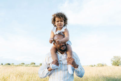 African american father covered eyes by cute little daughter on shoulders playing in field in summer and having fun looking away