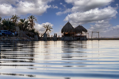 Stilt house on sea by palm trees and buildings against sky