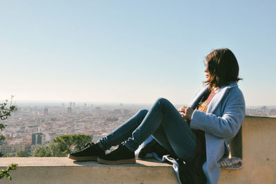 Woman sitting on retaining wall against clear sky