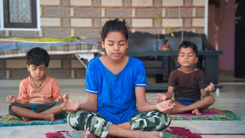 Three indian little kids doing meditate yoga asana on roll mat with eyes closed at home.