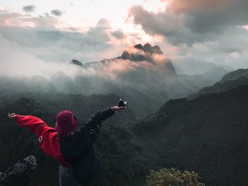 Rear view of woman with arms outstretched looking at mountains against cloudy sky during sunset