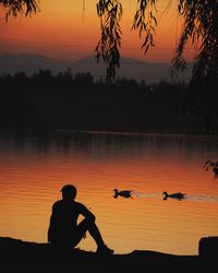 Silhouette man sitting by lake against sky during sunset