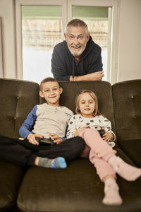 Girl and boy playing video game with grandfather leaning on sofa at home