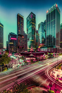 Light trails on city street by buildings against sky at night