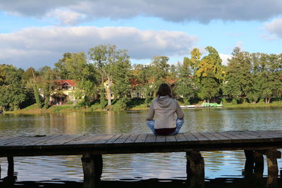 Rear view of woman sitting by lake against sky