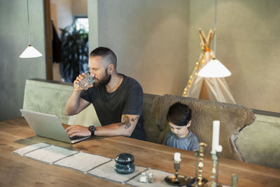 Man working on laptop while sitting with son at dining table