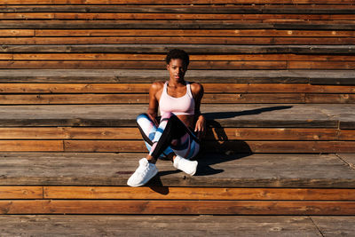 Slim african american young woman in colorful sport clothes and white sneakers looking at camera with curiosity and contemplating while sitting alone on brown wooden stairs on stadium