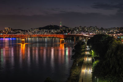 Illuminated bridge over river by buildings against sky at night