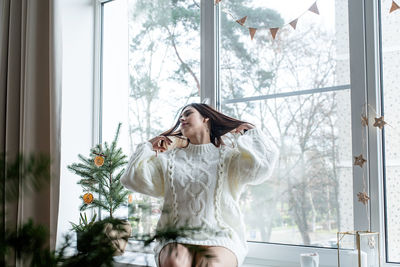Woman in warm white winter sweater sitting on the window at home at christmas eve holding cup