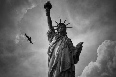 Low angle view of bird flying by statue against cloudy sky